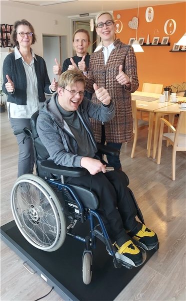 Drei Personen stehen hinter einer Rollstuhlwaage, darauf ist eine Person im Rollstuhl 