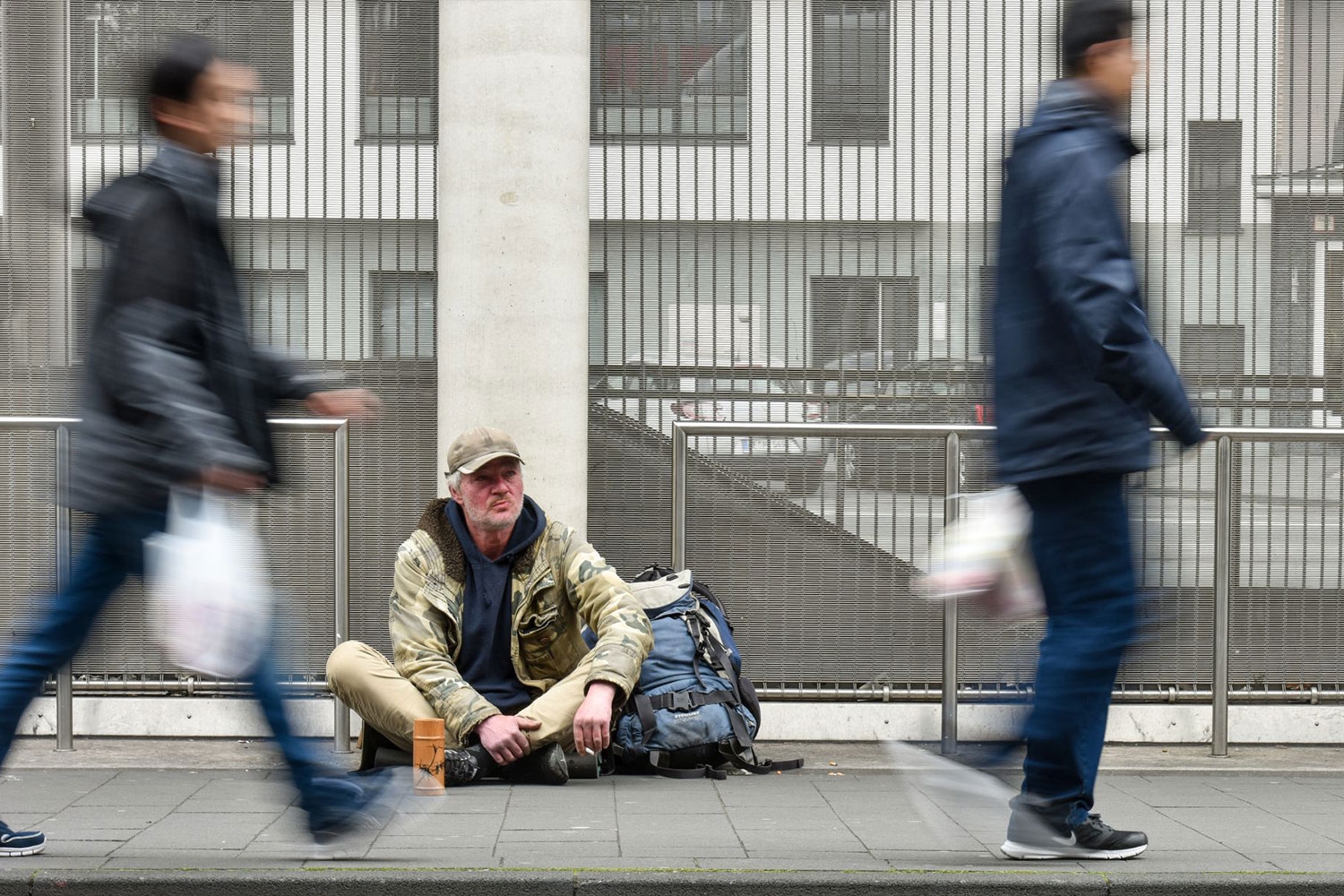 Ein obdachloser oder wohnungsloser Mensch sitzt auf der Straße. Passanten laufen vorbei.