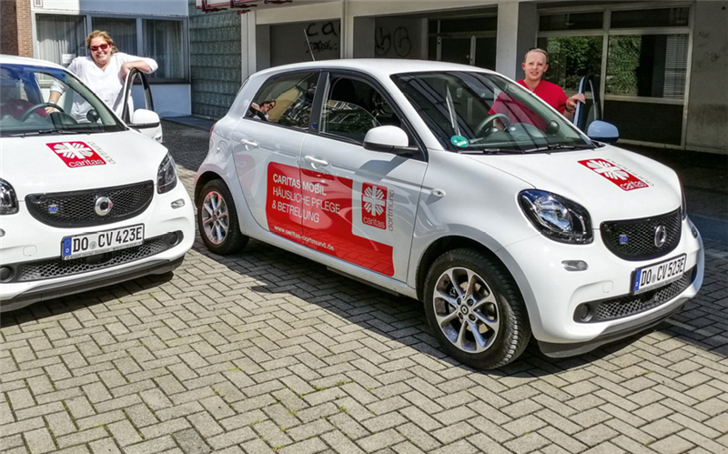 Zwei Pflegerinnen stehen neben zwei weißen Elektro-Autos des Caritasverbandes Dortmund
