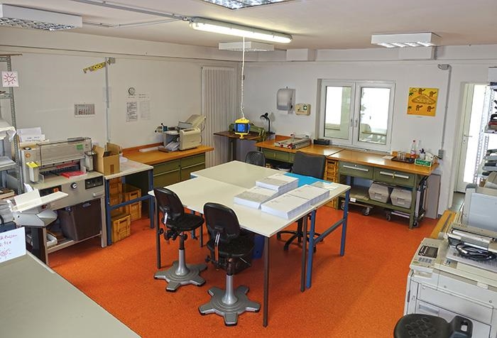 Arbeitsraum mit Papierbogen auf Tischen, sowie mehreren Maschinen für den Druckereigebrauch (Caritasverband Darmstadt e. V. / Jens Berger)