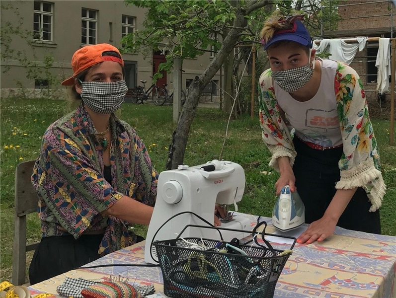 Zwei junge Frauen mit Mundschutz. Eine sitzt an einer Nähmaschine, die andere steht am Tisch mit einem Bügeleisen in der Hand.