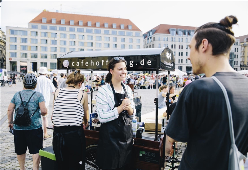 Auf dem Bild sind eine Mitarbeiterin sowie ein Besucher des „Café Hoffnung“ zu sehen, die sich neben dem mobilen Café unterhalten.