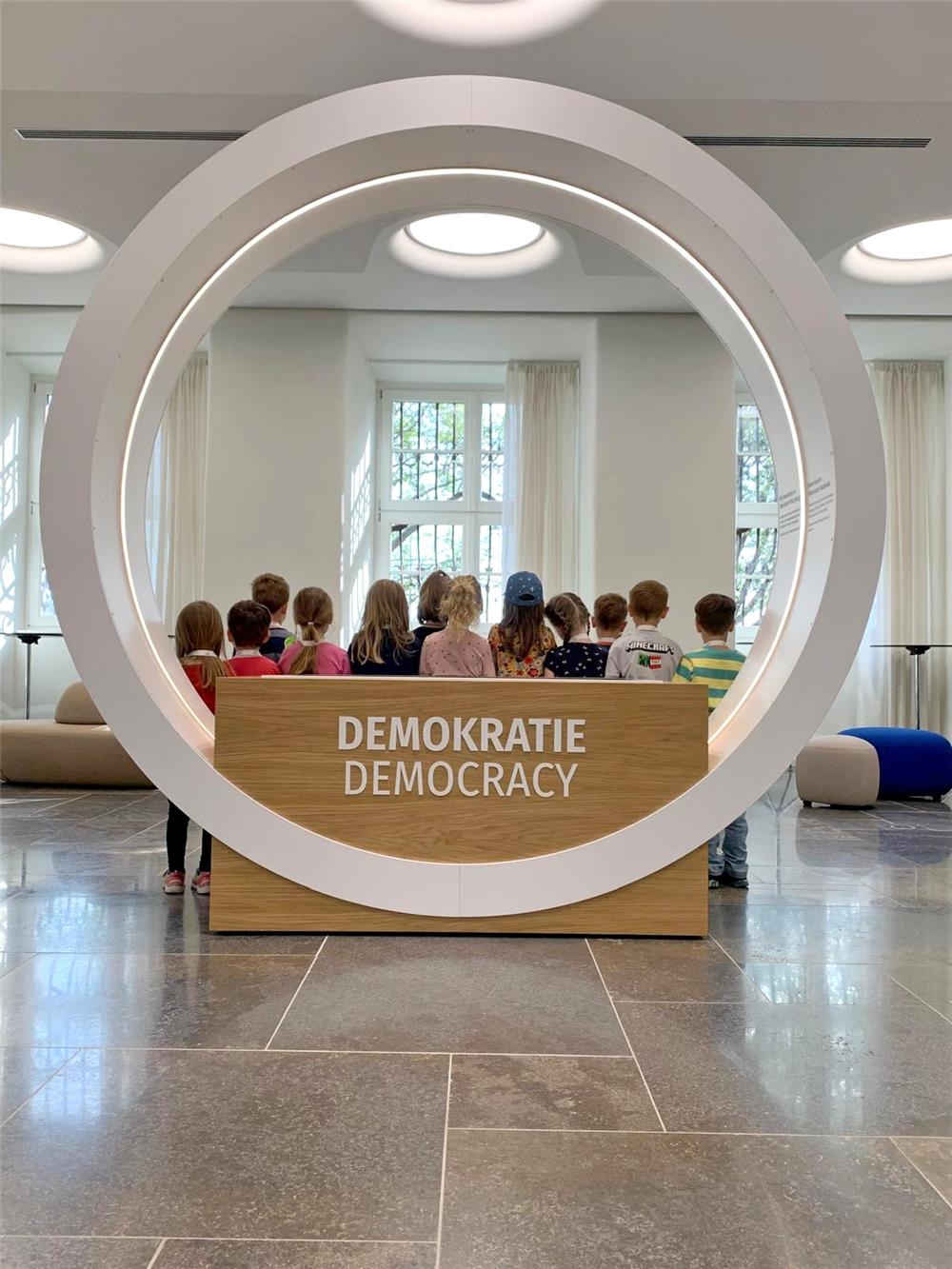 Kinder mit dem Rücken zur Kamera hinter einer Kreis-Skulptur mit der Aufschrift Demokratie