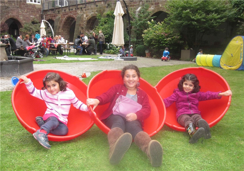 Im Hintergrund picknickende Erwachsene - im Vordergrund sitzen drei fröhliche Kinder in roten Spielschalen aus Kunststoff. (Susanne Leimbach)