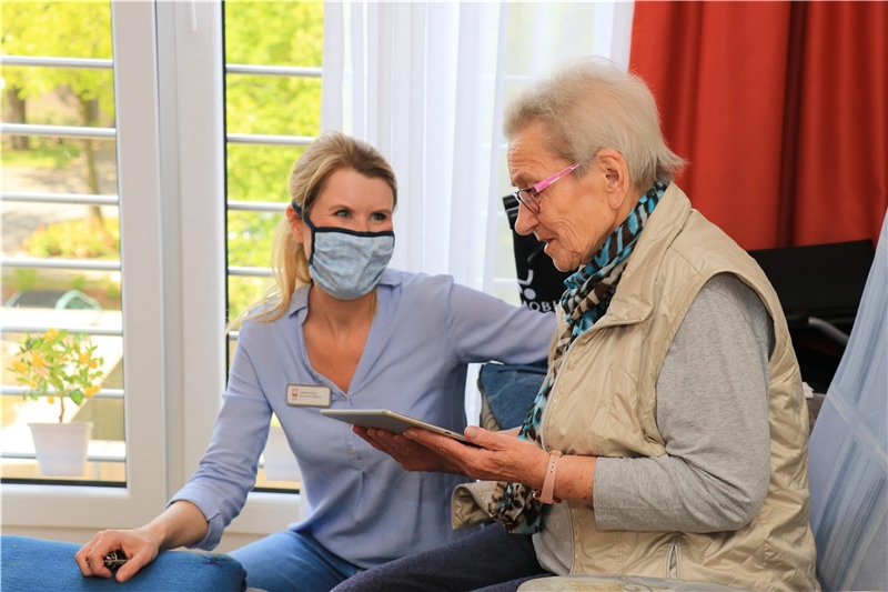 Eine Frau schaut eine ältere Dame an, die in einem Sessel sitzt und ein Tablet in ihren Händen hält.