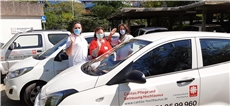 hinter einem weißen Auto der Caritas Pflege und Betreuung stehen drei Pflegekräfte mit Masken. Sie winken in die Kamera. Im Hintegrund auf der linken Seite des Gebäude der Caritas Pflege und Betreuung / (c) Caritasverband Hochtaunus