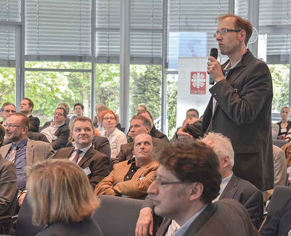 Das Publikum des 2. Kirchlichen Dienstgebertags in Duisburg, das auf Stühlen sitzt und einem stehenden Zuschauer mit Mikrofon zuhören. (Michael Kreuzfelder)