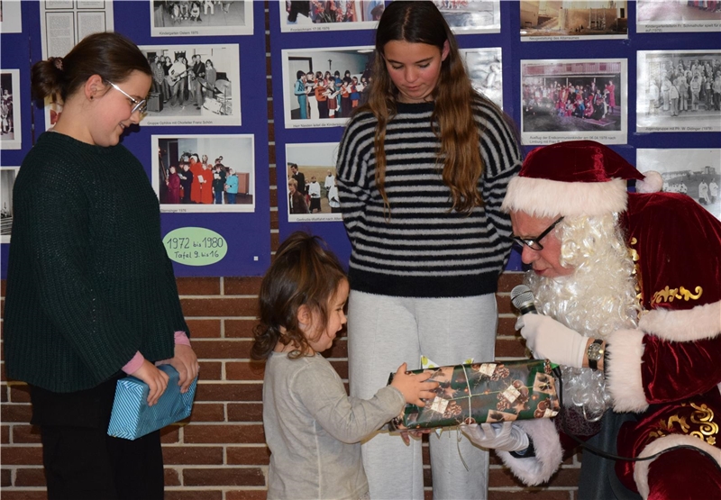 Ein Weihnachtsmann überreicht einem kleinen Kind ein Geschenk, während zwei ältere Mädchen daneben stehen. Im Hintergrund sind Fotos und blaue Plakate zu sehen.