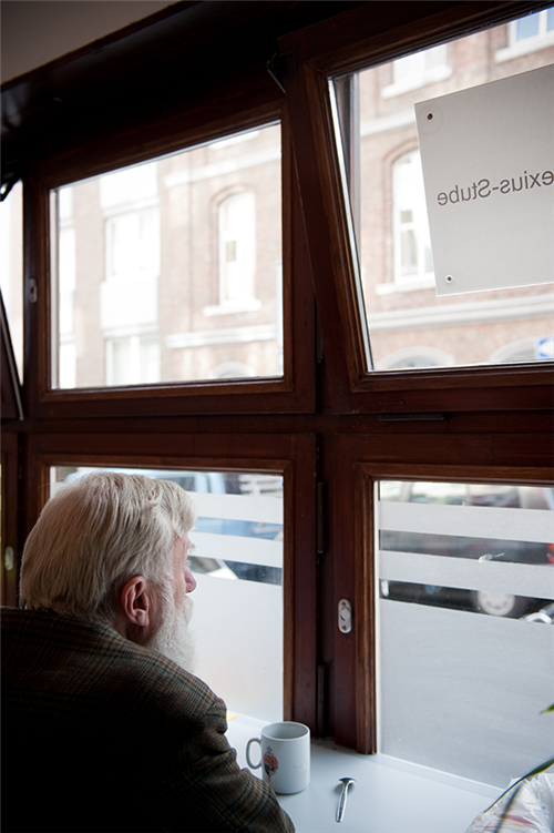 Ein älterer Mann sitzt, mit dem Rücken zur Kamera gewandt, an einer Fensterfront der St.-Alexius-Stube und blickt hinaus. Vor ihm steht eine Tasse mit Löffel. (Caritas)