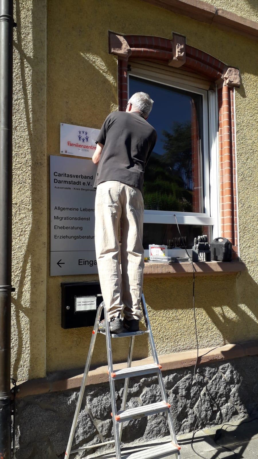 Ein Mann steht auf einer Leiter und befestigt ein Schild. (Caritasverband Darmstadt e. V.)