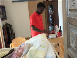 Ein junger Pfleger steht am Bett einer alten Frau und gibt ihr mit einem Löffel zu Essen / Foto: Schedlbauer