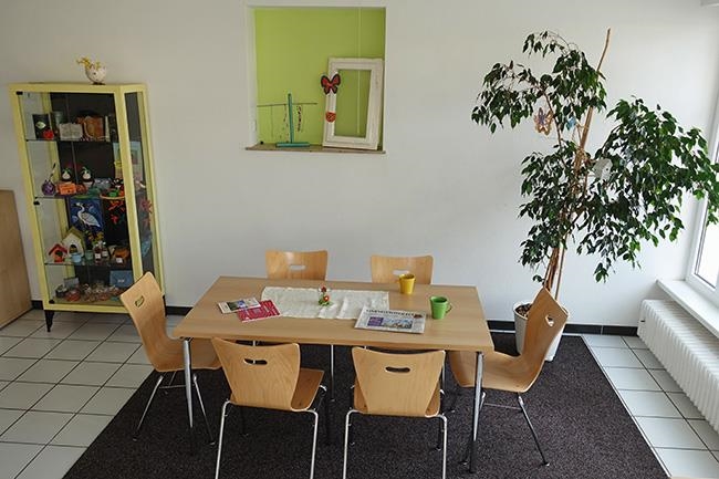 Tisch mit sechs Stühlen, darauf Kaffeetassen und Zeitungen, ein Benjamini und ein Schrank mit Kunstgewerblichen Artikeln darin (Caritasverband Darmstadt e. V. / Jens Berger)
