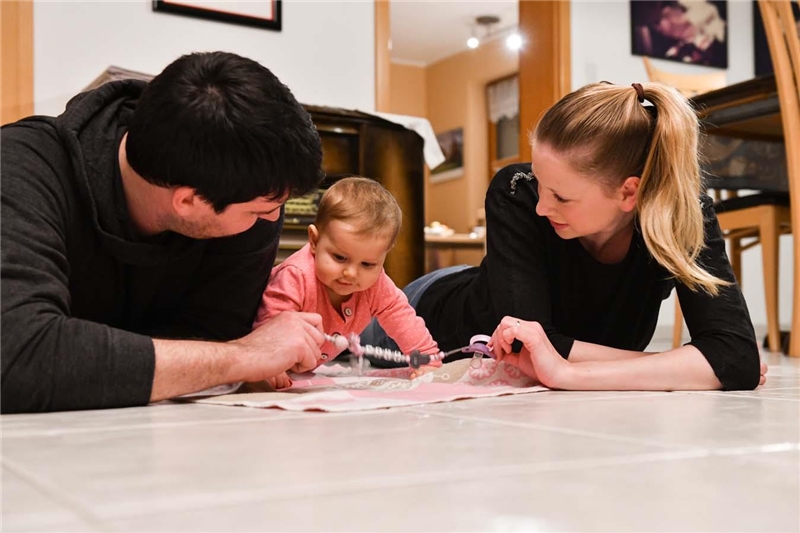 Eine junge Familie mit Kleinkind liegt auf dem Boden. Die Eltern sehen das Baby liebevoll an.