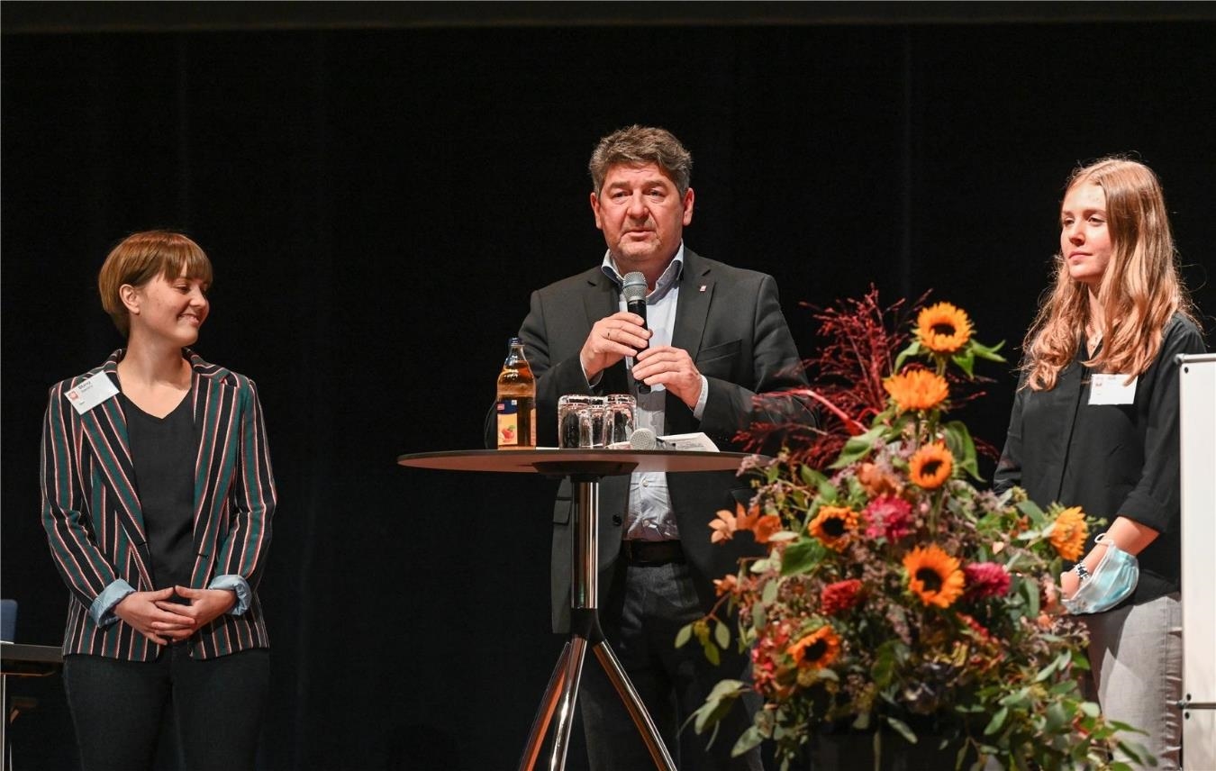 Drei Menschen stehen um einen runden Tisch auf der Bühne. Mann in der Mitte hält Mikrofon in der Hand.  (Deutscher Caritasverband / Harald Oppitz)