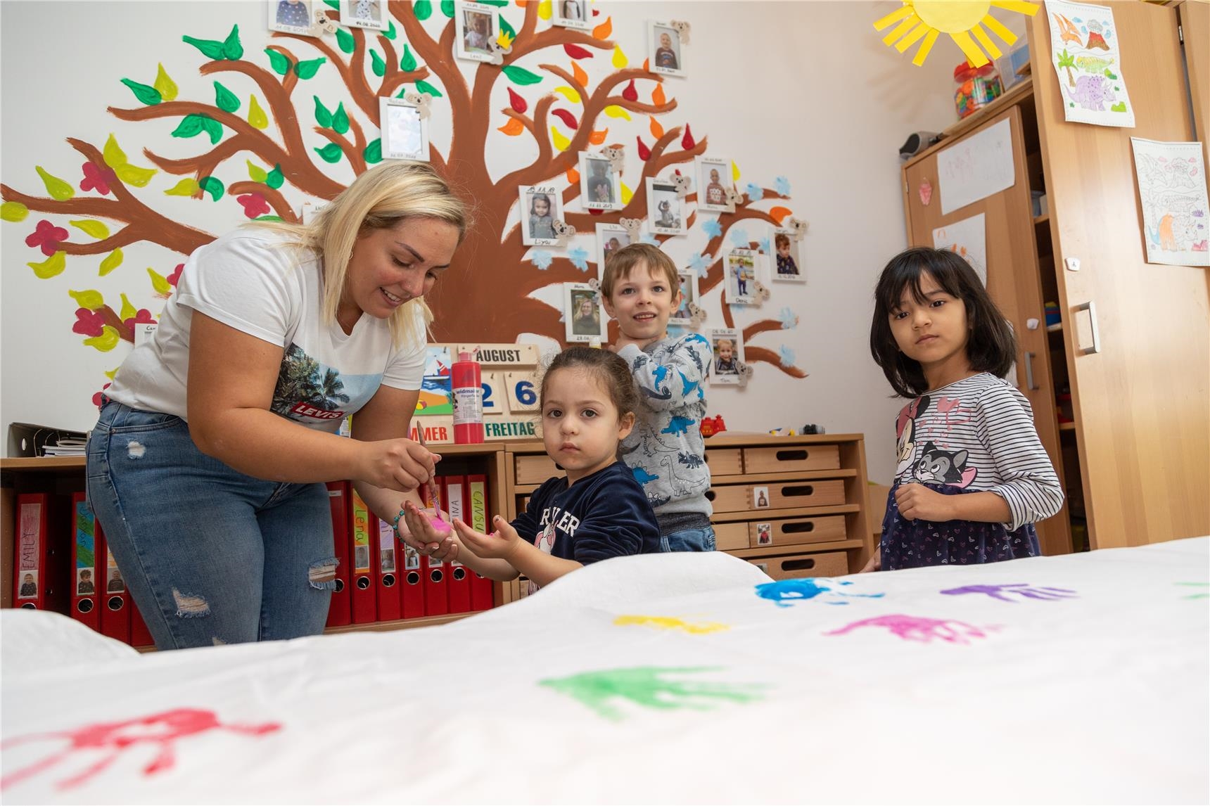 Eine Erzieherin bemalt die Hände der Kinder mit Fingermalfarben. 