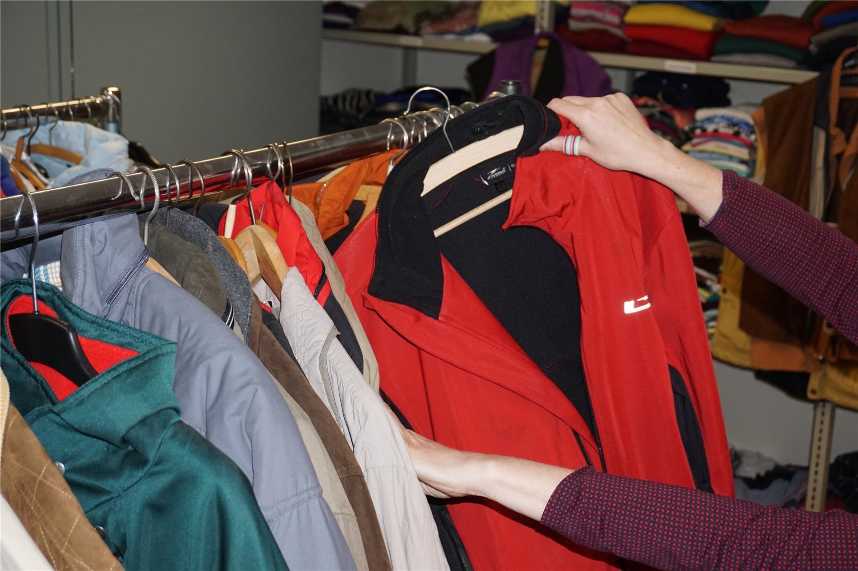 Zwei Hände halten eine Jacke, die auf einem Kleiderständer mit vielen Jacken in verschiedenen Farben hängt (Jutta Link)