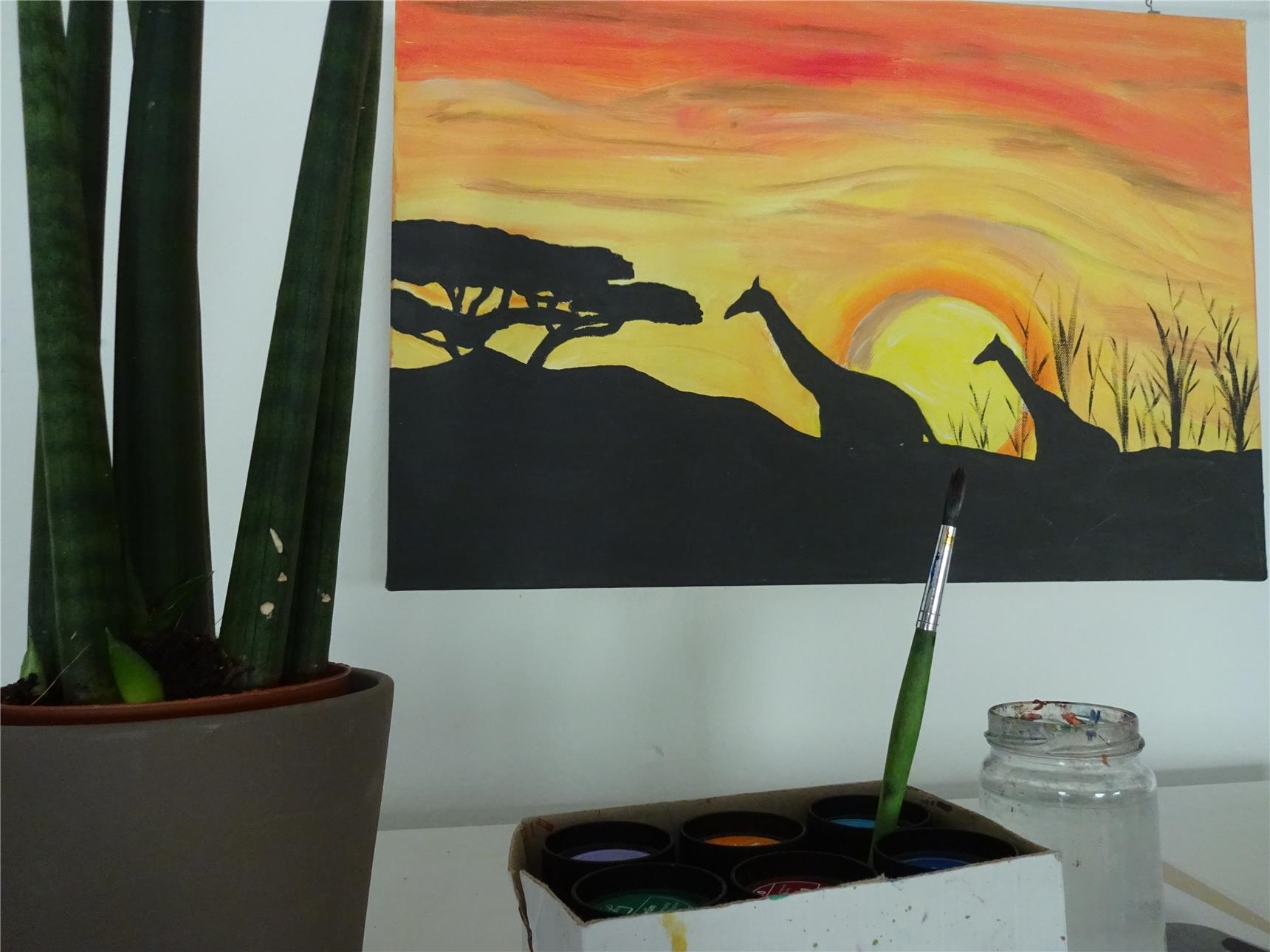 An einer Wand hängt ein Bild mit Silhouetten von Giraffen vor einem Sonnenuntergang; davor: Farben, ein Pinsel und ein Wasserglas. (Tagesstätte St. Maximin/Caritasverband Trier e. V.)