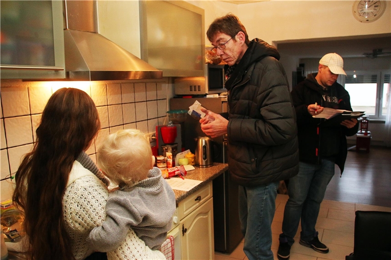 Das Foto zeigt zwei Männer in einer Küche, die mit einer Frau mit Kind auf dem Arm sprechen.