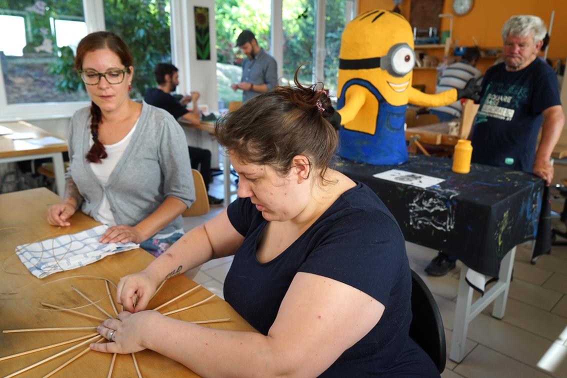 Eine Frau bearbeitet an einem Tisch einen Korbrohling, eine andere schaut zu. (Caritasverband Trier e. V.)
