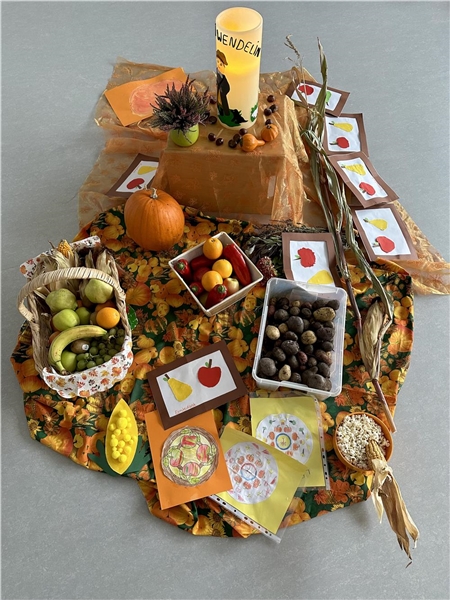 Auf dem Boden stehen herbstliche Dekoration, ein Korb mit Obst und weiteren Gaben rund um das Erntedankfest, in der Mitte steht eine Kerze