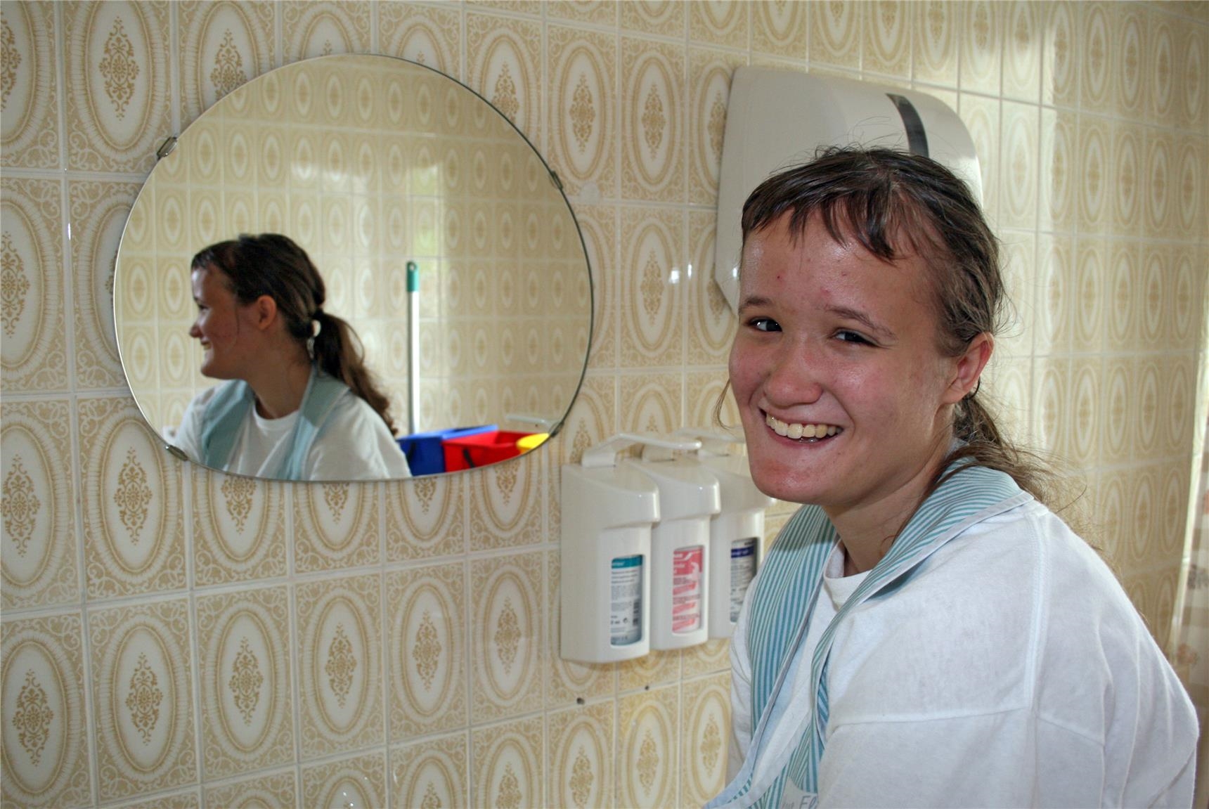 Eine junge Frau in Berufskleidung steht in einem Badezimmer vor einem Spiegel und lacht. (BBZ Haus Elisabeth/Caritasverband Trier)