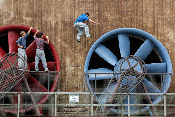 Zwei Jugendliche stehen auf Gittern an einem großen roten Ventilator und blicken auf einen Jungen, der von dem roten auf eine daneben gelegene blauen Ventilator springt (Foto: Achim Pohl)