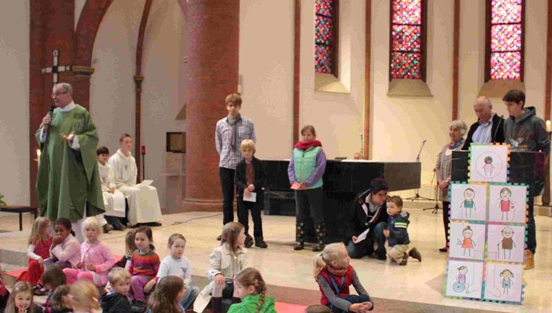 Kinder, der Pfarrer und weitere Gemeindemitglieder im Altarraum. Über Mikrofon werden Erläuterungen zu den Kinderbildern gegeben. (Manuela Siegerink-Schröder)