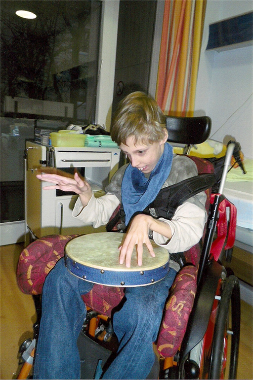 Ein Junge mit Behinderung der in einem Rollstuhl sitzt und auf einem Tambourin spielt. Der Rollstuhl steht in einem Krankenzimmer. (Caritas)