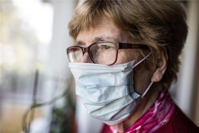 Seniorin mit Mund-Nase-Schutz schaut aus dem Fenster