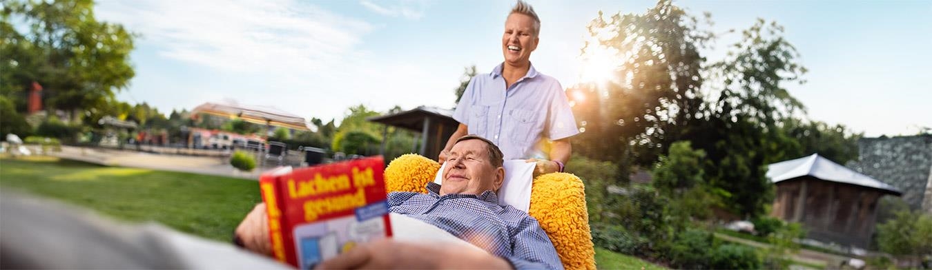 Ein Mann liegt in einem Rollstuhl im Freien und liest. Hinter ihm steht eine Pflegekraft.