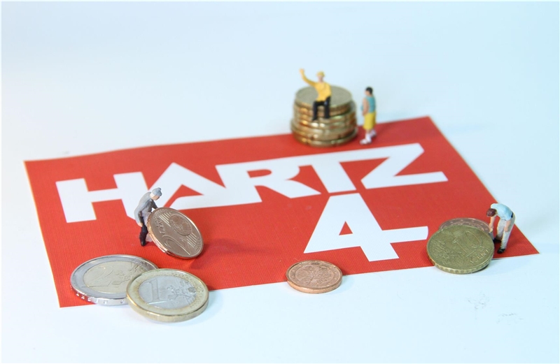 Die Grafik zeigt auf einer Unterlage mit dem Schriftzug "Hartz IV" Spielfiguren rund um Geldmünzen.