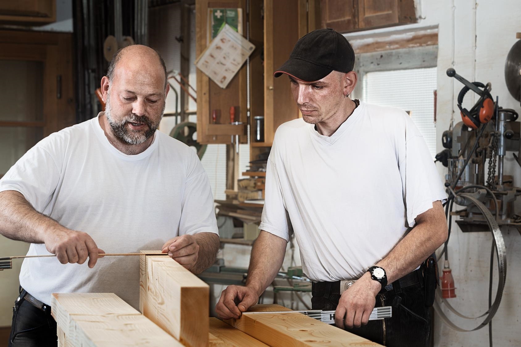 Zwei Männer mit Zollstöcken und Holzbalken in einer Werkstatt.