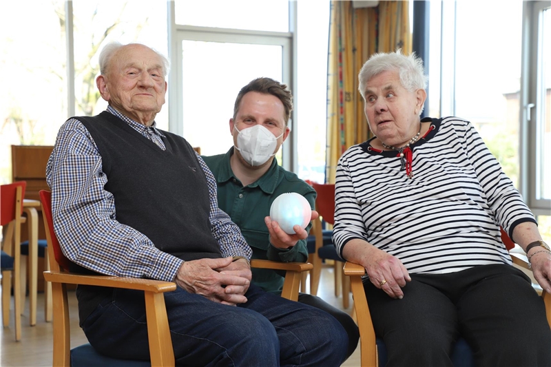 Das Foto zeifgt einen jungen Mann zwischen einem alten Mann und einer alten Frau, der einen Ball in der Hand hält.