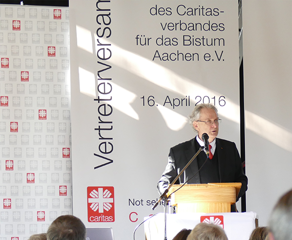 Prof. em. Klaus J. Bade steht an einem Pult und hält eine Rede. Im Vordergrund sind einige Teilnehmer der Vertreterversammlung des DiCV Aachen zu sehen.