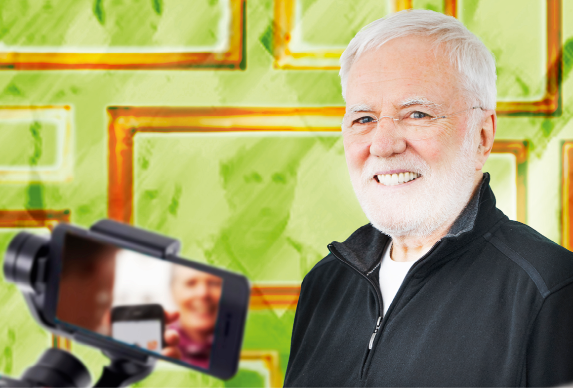 Porträt: Gerd Fischer, einem hell-grünen Hintergrund mit goldenen Bilderrahmen. Im Vordergrund ist ein Smartphone auf einem Selfie-Stick zu sehen.