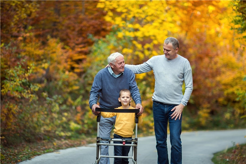 Ein älterer Mann mit Rollator geht auf einem Waldweg spazieren. Vor ihm geht sein Enkel. Neben ihm sein erwachsener Sohn der ihm einen Arm auf die Schulter legt.