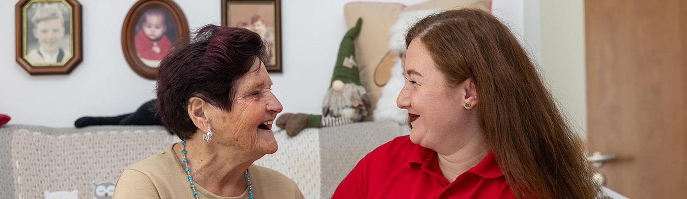 Eine Pflegekraft und eine Seniorin sitzen auf einem Sofa und lächeln sich an.