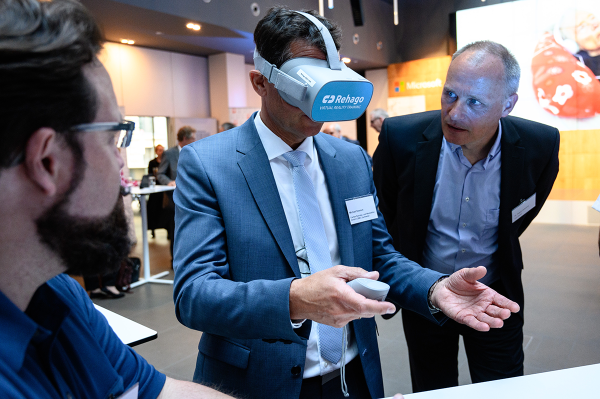 Michael Doersch und Jürgen Holtkamp stehen mit einem Mann am Stand zum Virtual Reality Training Rehago. Michael Doersch trägt dabei eine VR-Brille. (Charles Yunck)