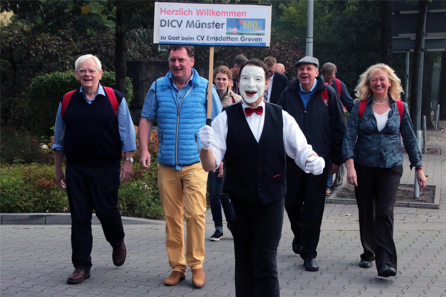 Das Foto zeigt eine wandernde Gruppe eine Straße überquerend mit einem Pantomimen vorneweg, der ein Schild in die Höhe hält. (Harald Westbeld)