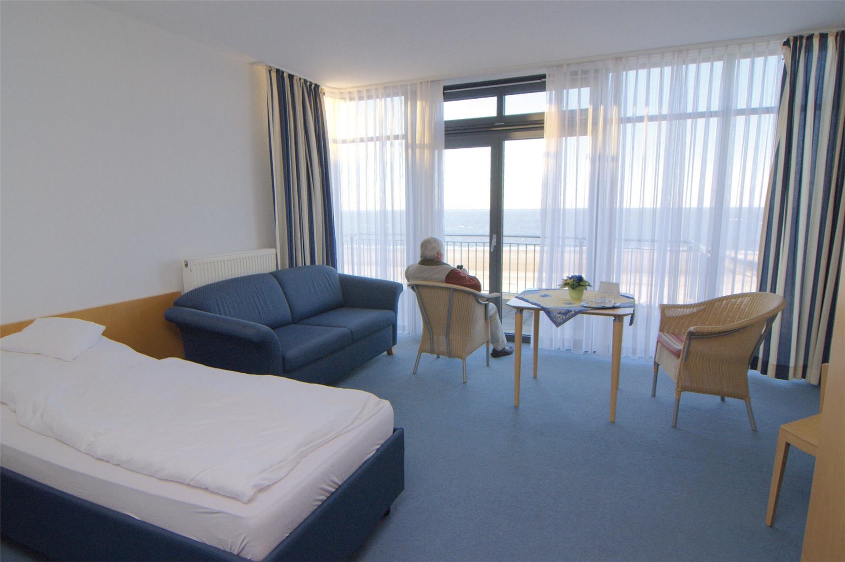 Ein Gästezimmer mit Bett, Sofa, Sitzecke und großem Panoramafenster mit Meerblick. 