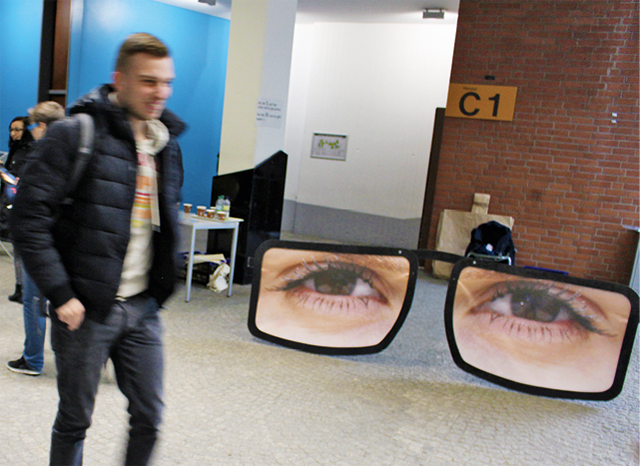 Ein junger Mann geht in den Räumen der KatHo Paderborn an einer großen, schwarzen Brille mit Augen vorbei, die dort aufgestellt wurde (youngcaritas)