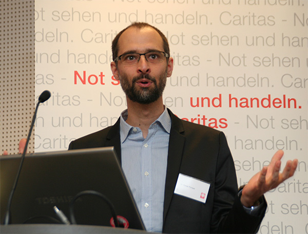 Dr. Florian Pomper steht gestikulierend vor einem aufgeklappten Laptop an einem Pult und hält eine Rede. Im Hintergrund ist ein Caritas-Rollup zu sehen. (Harald Westbeld)