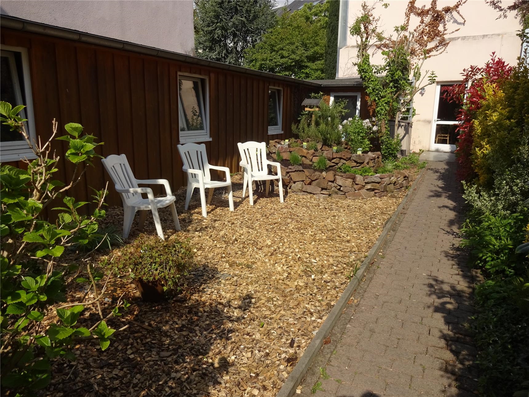 Drei Stühle in einem Hof auf einem kleinen mit Rindenmulch bedeckten Platz, links und rechts Büsche, daneben ein Kräuterbeet. (Tagesstätte St. Maximin/Caritasverband Trier e. V.)