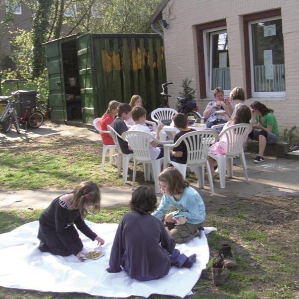 Außenbereich der Marienburg bei schönem Wetter, mit zwei auf einer Decke spielenden Kindern im Vordergrund und mehreren Kindern und Erwachsenen im Hintergrund, an einem Tisch 