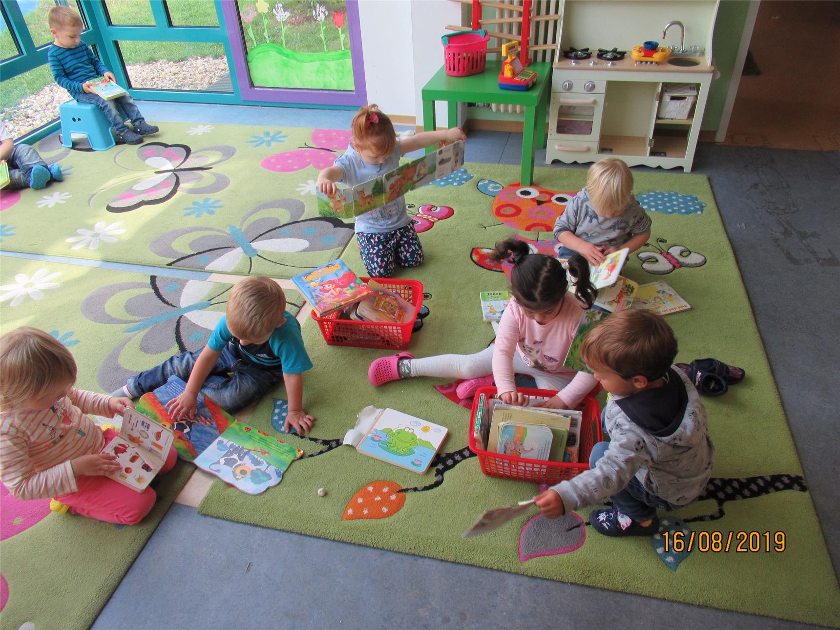 Mehrere Kinder sitzen auf bunten Teppichen und schauen Bilderbücher an 