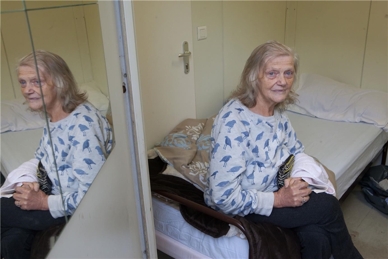 ältere Frau sitzt auf ihrem Bett im Wohncontainer und schaut aus der Tür