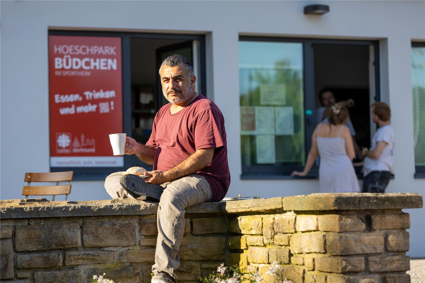 Ein Mann sitzt auf einer Mauer und trinkt einen Kaffee. 