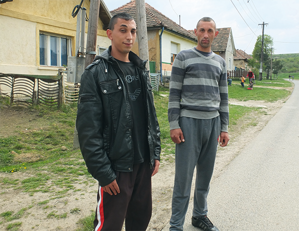 Zwei junge Männer stehen in einem ländlichen Gebiet vor einigen Wohnhäusern an einer Straße und blicken in die Kamera (Christoph Grätz)