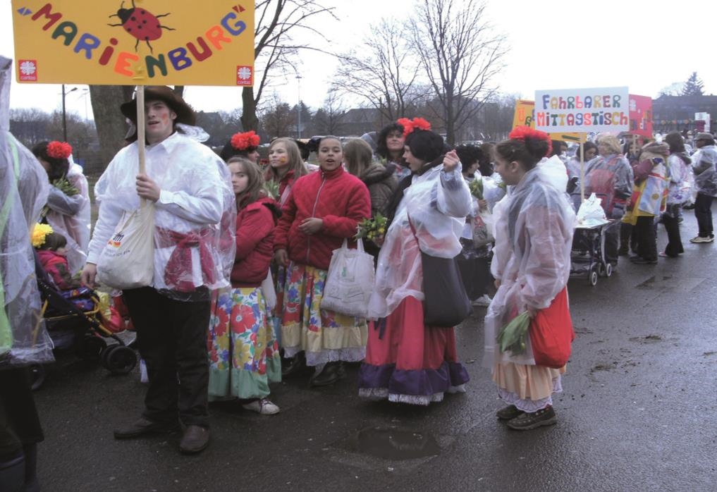 Eine Gruppe der Marienburg bei einem Karnevalszug, im Vordergrund ein Mann mit einem Marienburg-Schild, es regnet 