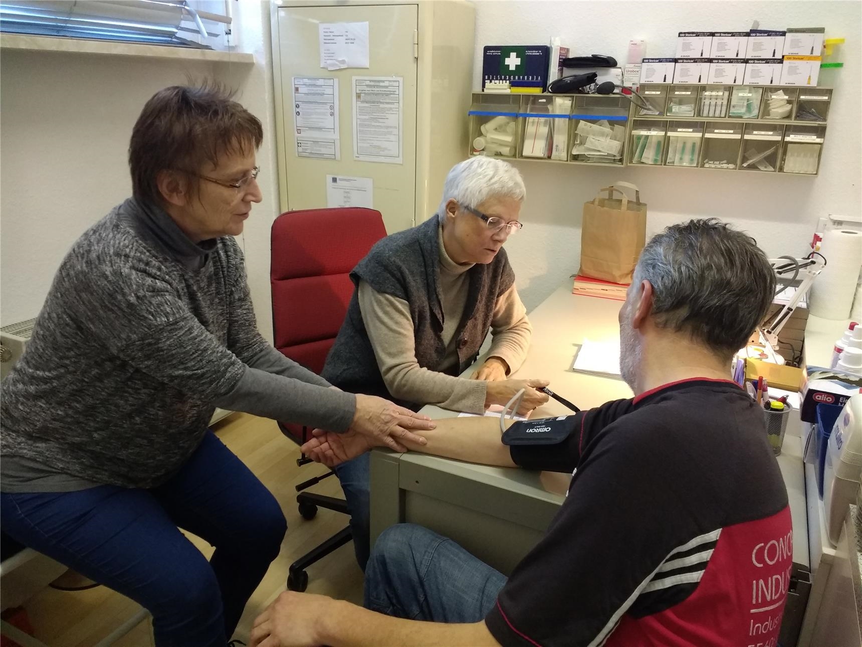 Zwei Frauen sitzen an einem Tisch gegenüber von einem Mann, der Blutdruck gemessen bekommt.  (Foto: Caritasverband Worms)